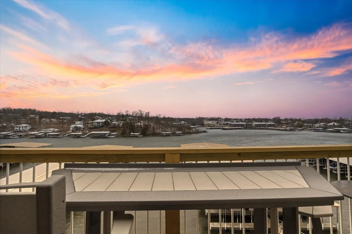 The hero image captures a serene lakeside sunset as viewed from the condo, with the soft glow of the sky reflected on calm waters. In the distance, lakeside homes dot the opposite shore, and a spacious outdoor area is visible—perfect for dining, relaxing, and taking in the views. The scene evokes a tranquil, inviting atmosphere ideal for gatherings and unwinding.