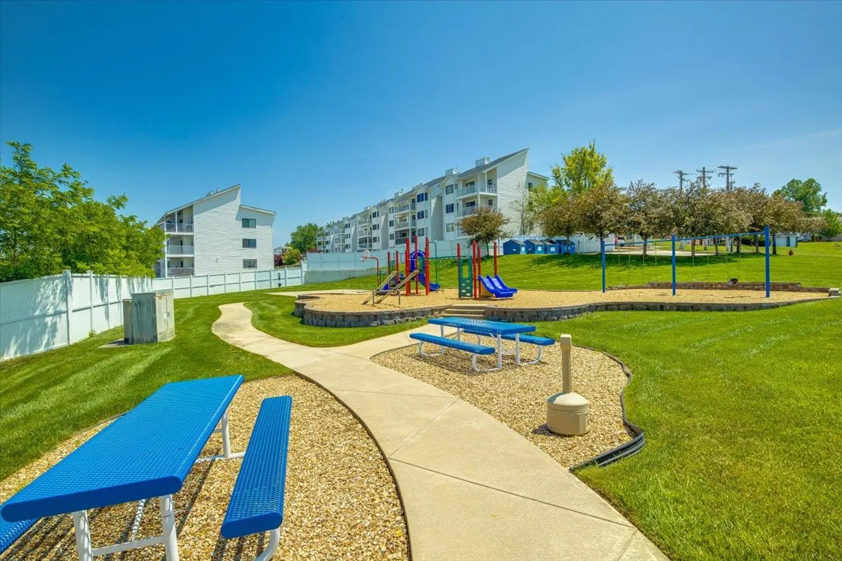 A joyful scene of a playground with colorful equipment, benches, and a green lawn stretching in front of apartment-style resort buildings. Children can be seen playing, while adults relax nearby, evoking a family-friendly environment.