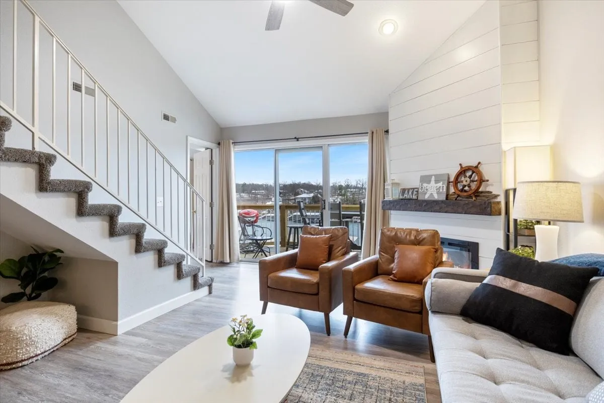 A cozy living room featuring a large sofa and chairs arranged around a central coffee table, with a fireplace at one end and expansive windows showcasing serene lake views. The space is bathed in natural light, and the setup invites both relaxation and group activities like game nights.