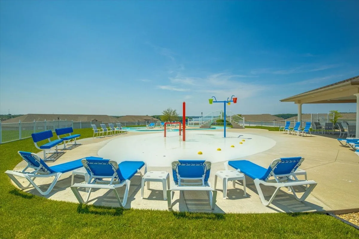 A lively splash pad with colorful water features, lounge chairs, and a green lawn. Children are actively engaged in playful water activities, while adults relax nearby, creating a vibrant family-friendly atmosphere.