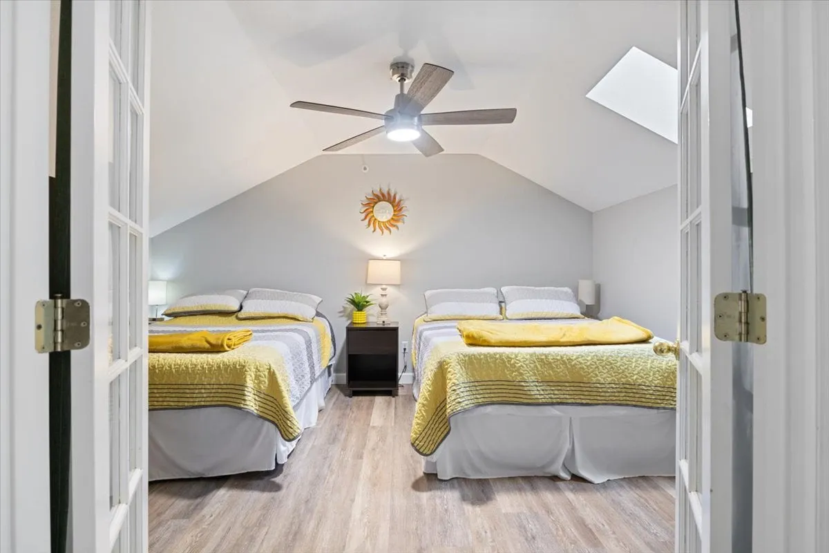 A cheerful room featuring two beds with yellow blankets, a ceiling fan overhead, sun-themed wall decor, and a nightstand with a lamp and plant. The layout suggests a flexible space that could serve families or groups, ideal for late-night chats or relaxed mornings.