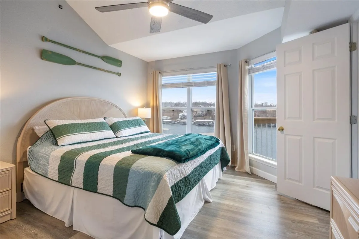 This image features a cozy bedroom with a striped quilt on the bed, large windows letting in natural light, and decorative paddles on the wall. The view out the windows hints at a scenic outdoor environment, and the soft textures and calm color palette evoke serenity and comfort.