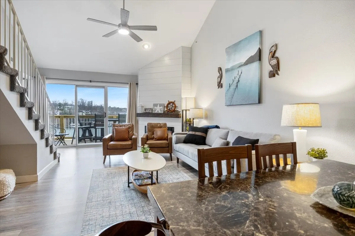 A bright living-dining space with a dining table, sofa, and artwork, all oriented toward a private balcony with a scenic view. The balcony is visible, suggesting access to outdoor dining and lakeside relaxation.
